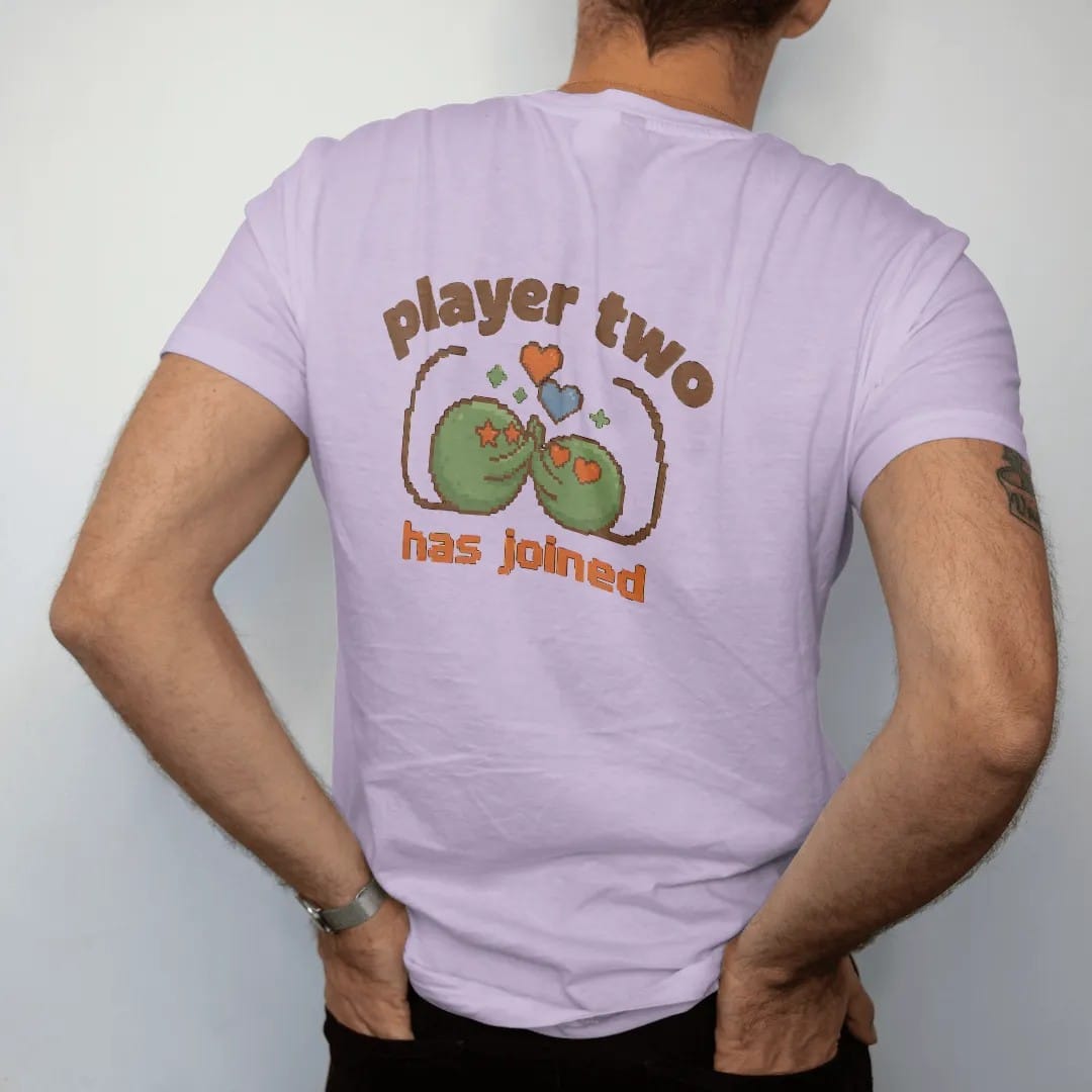 Man wearing a Orchid tee, seen from the back: two pixel slimes high-fiving under pixel hearts with controller cords curling into a heart, under player two in arcade letters and has joined in pixel type, in brown, rust, moss and denim.