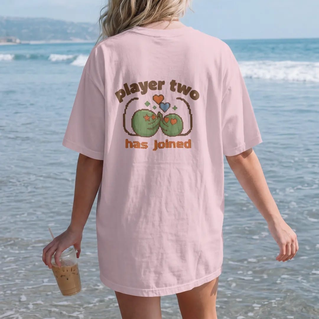 Woman on the beach in a Blossom tee, back view: two pixel slimes high-fiving under pixel hearts with controller cords curling into a heart, under player two in arcade letters and has joined in pixel type, in brown, rust, moss and denim.
