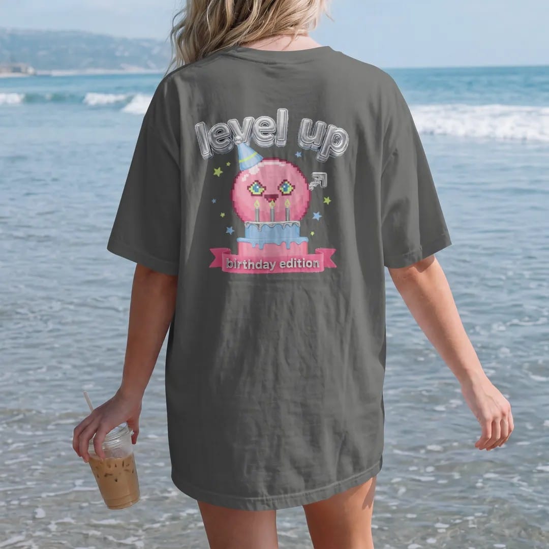 Woman on the beach in a Pepper tee, back view: blip the pixel slime in a party hat holding a pixel birthday cake with confetti stars, under level up in chrome bubble letters and a birthday edition ribbon, in pink, lime, blue and silver.