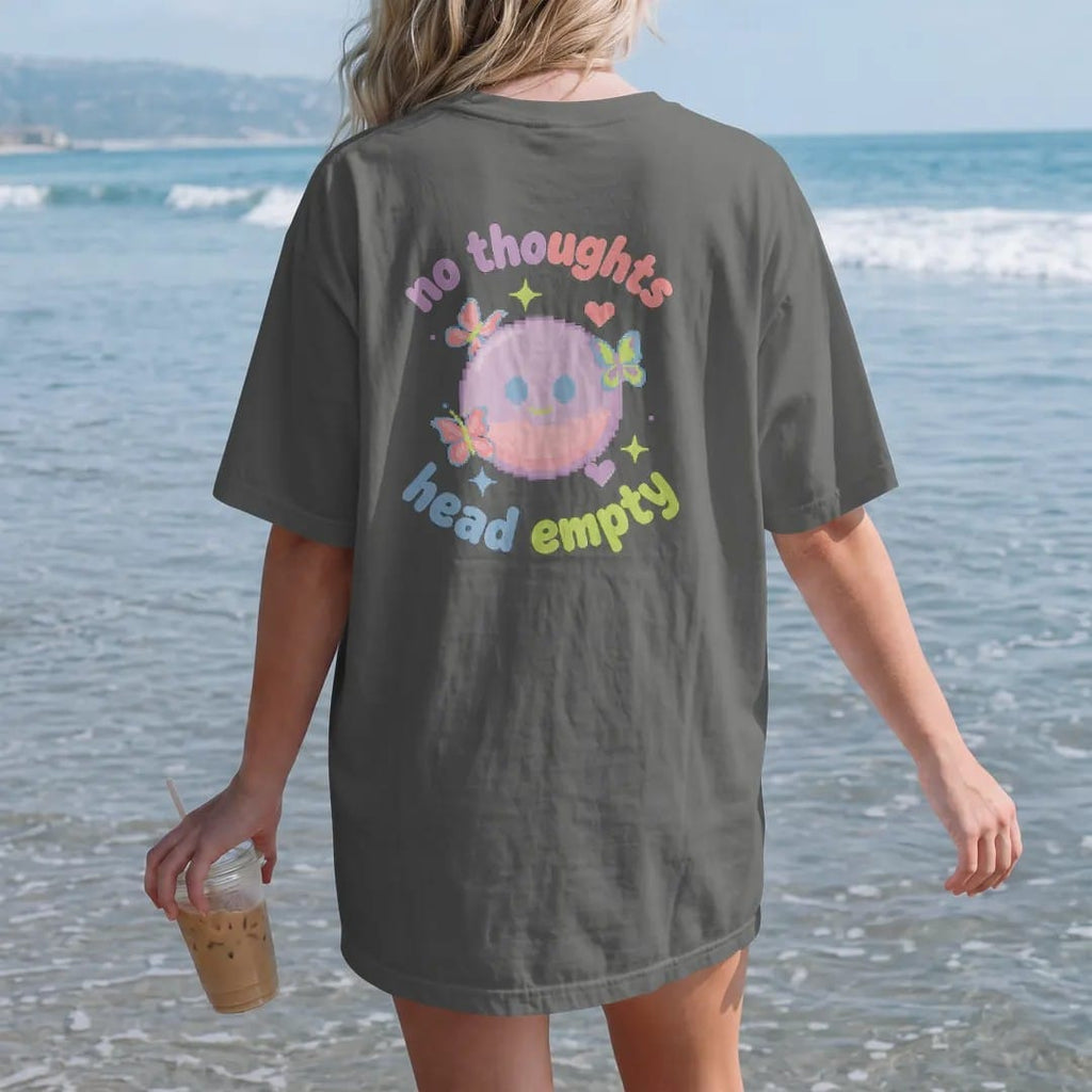 Woman on the beach in a Pepper tee, back view: blip the pixel slime with blank dot eyes floating among butterflies, stars and hearts, between no thoughts and head empty in lilac, pink and blue wavy bubble letters.