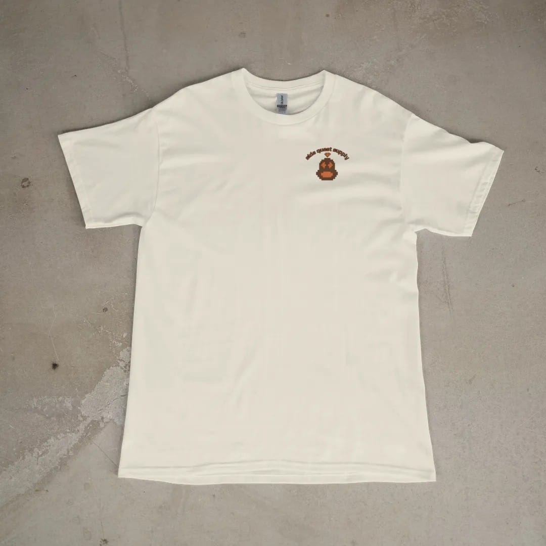 Front of a Ivory tee laid flat, with the small Side Quest Supply logo on the left chest.