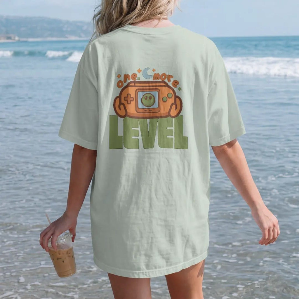 Woman on the beach in a Bay tee, back view: a generic retro handheld console showing Blip the pixel slime, held under a moon and stars, with one more in arcade letters and level in big block type, in brown, rust, moss and denim.