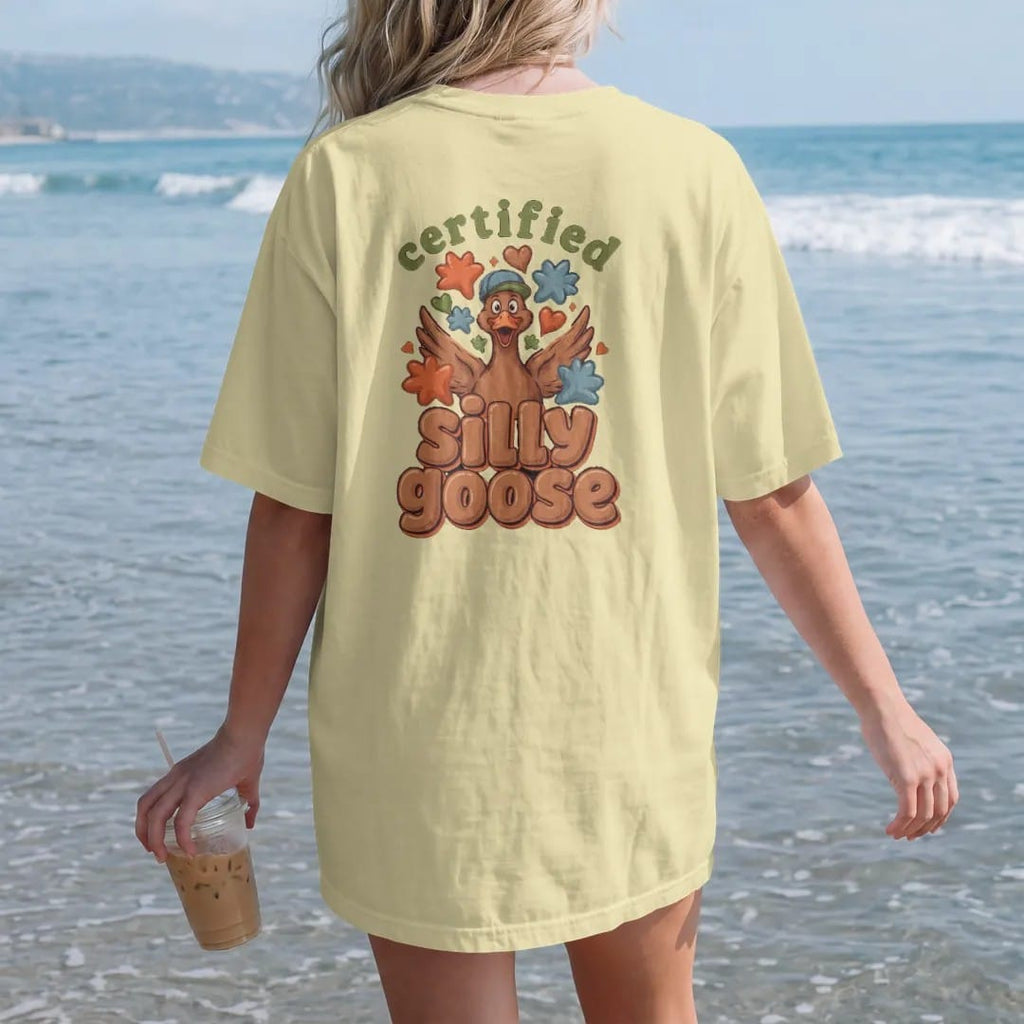 Woman on the beach in a Butter tee, back view: a goofy goose in a backwards cap surrounded by puffy stars and hearts, with certified in an arched stamp and silly goose in fat bubble letters, in brown, rust, moss and denim.