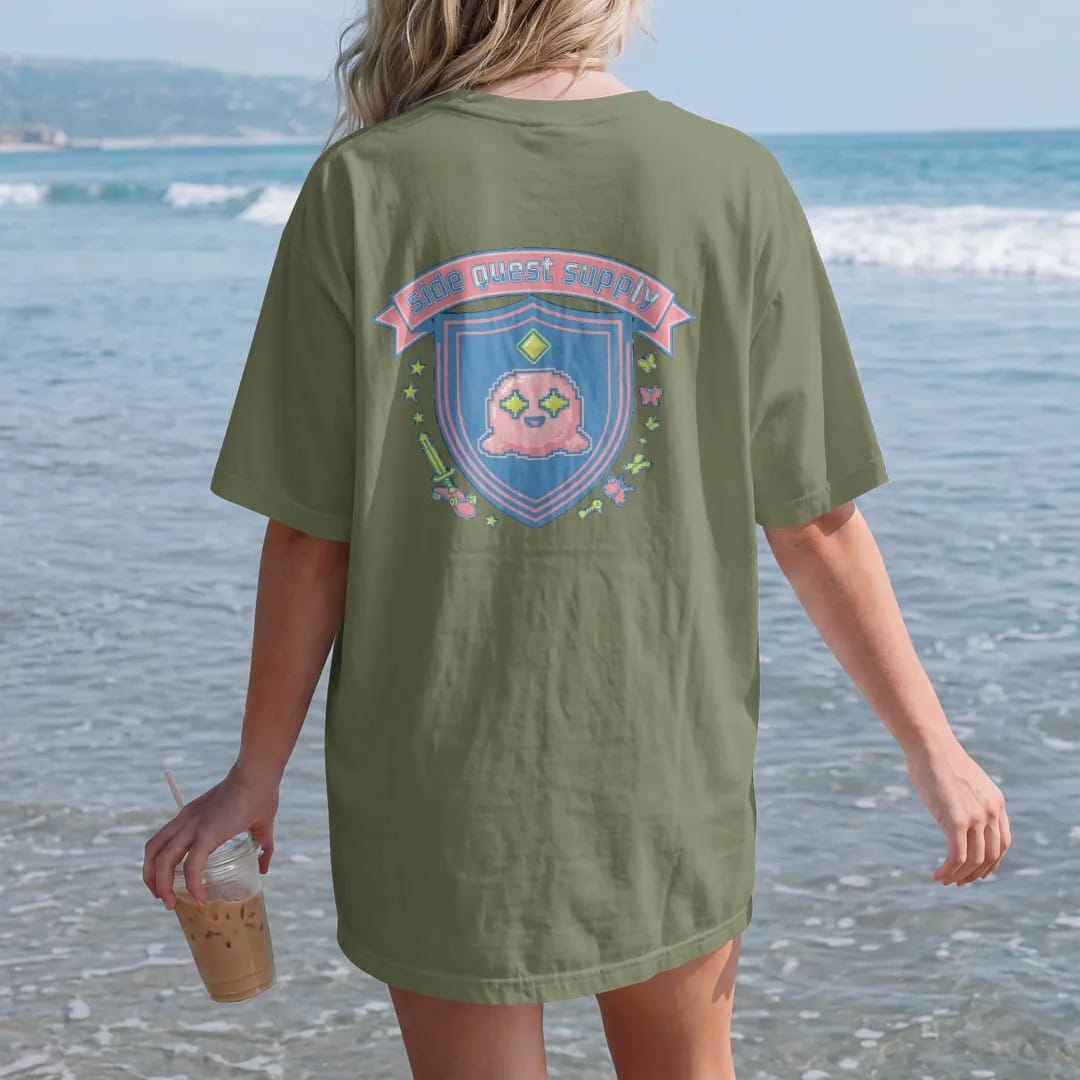 Woman on the beach in a Moss tee, back view: a varsity crest with Blip the pixel slime under a quest marker, a crossed skateboard and pixel sword, with side quest supply in an arched varsity banner, in pink, lime, yellow and blue.