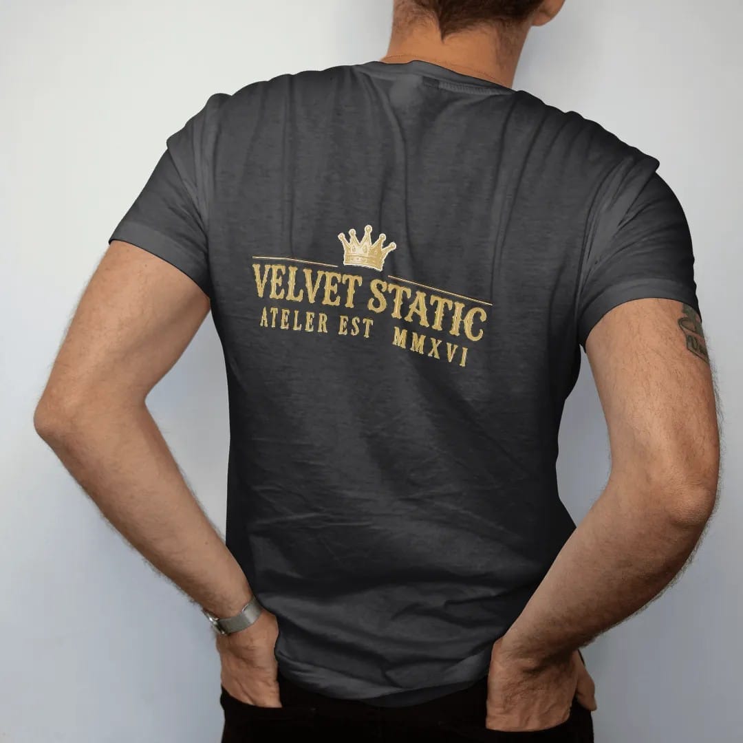 Man wearing a Black tee, seen from the back: a fictional house lockup with Velvet Static in large gothic blackletter between thin rules and Atelier Est. MMXXVI in spaced serif capitals under a small crown, in bone cream and antique gold.