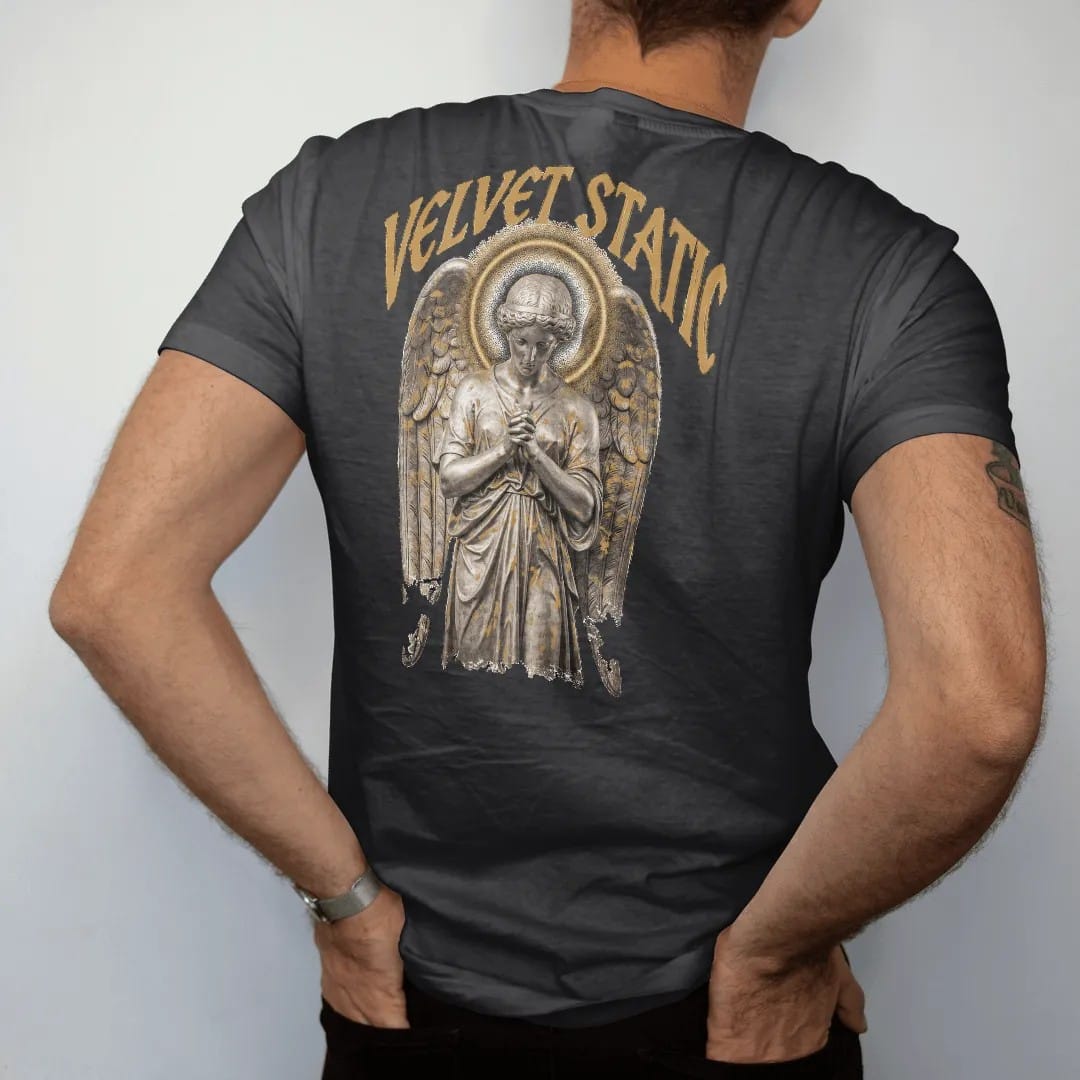 Man wearing a Black tee, seen from the back: a classical angel with folded wings and a halo of TV static in airbrushed chrome, under Velvet Static in tall gothic blackletter, in bone cream, chrome silver and antique gold.