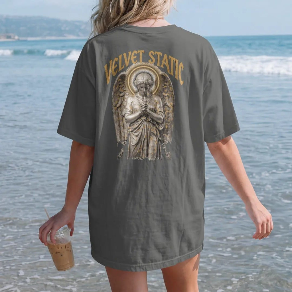Woman on the beach in a Pepper tee, back view: a classical angel with folded wings and a halo of TV static in airbrushed chrome, under Velvet Static in tall gothic blackletter, in bone cream, chrome silver and antique gold.