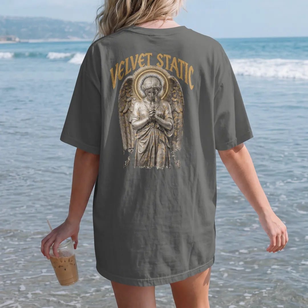 Woman on the beach in a Pepper tee, back view: a classical angel with folded wings and a halo of TV static in airbrushed chrome, under Velvet Static in tall gothic blackletter, in bone cream, chrome silver and antique gold.
