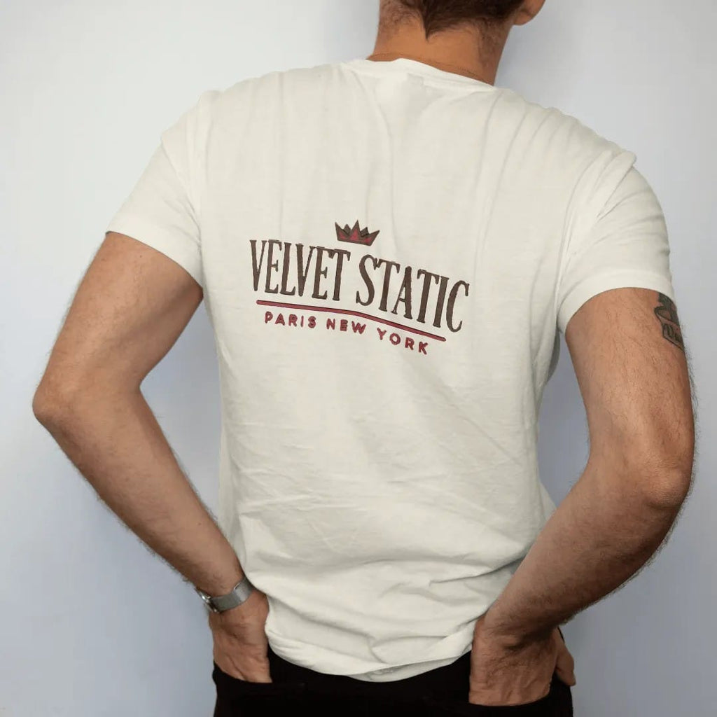 Man wearing a Ivory tee, seen from the back: a clean fashion-house lockup with Velvet Static in a large spaced didone serif over a thin rule and Paris New York in widely spaced sans capitals, under a small crown, in espresso and oxblood.