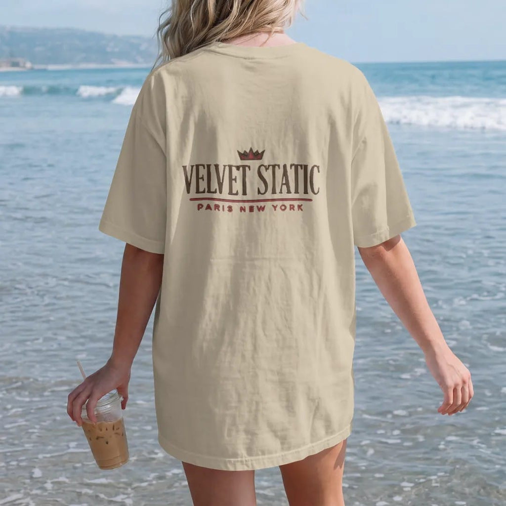 Woman on the beach in a Sandstone tee, back view: a clean fashion-house lockup with Velvet Static in a large spaced didone serif over a thin rule and Paris New York in widely spaced sans capitals, under a small crown, in espresso and oxblood.