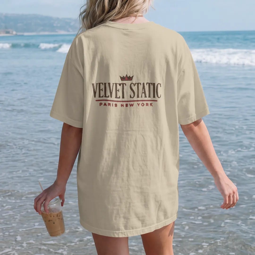 Woman on the beach in a Sandstone tee, back view: a clean fashion-house lockup with Velvet Static in a large spaced didone serif over a thin rule and Paris New York in widely spaced sans capitals, under a small crown, in espresso and oxblood.