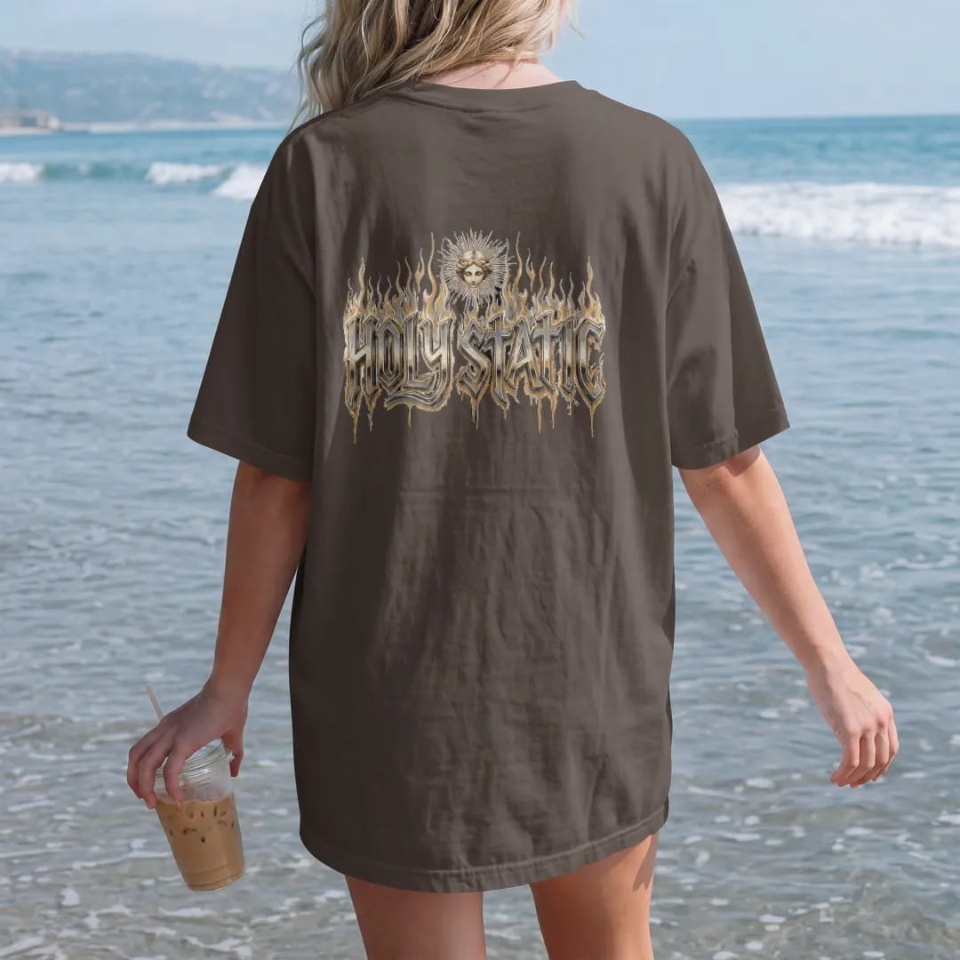 Woman on the beach in a Espresso tee, back view: Holy Static in huge liquid-chrome gothic blackletter with tall flames behind it and a small static-haloed angel head above, in bone cream, chrome silver and antique gold.
