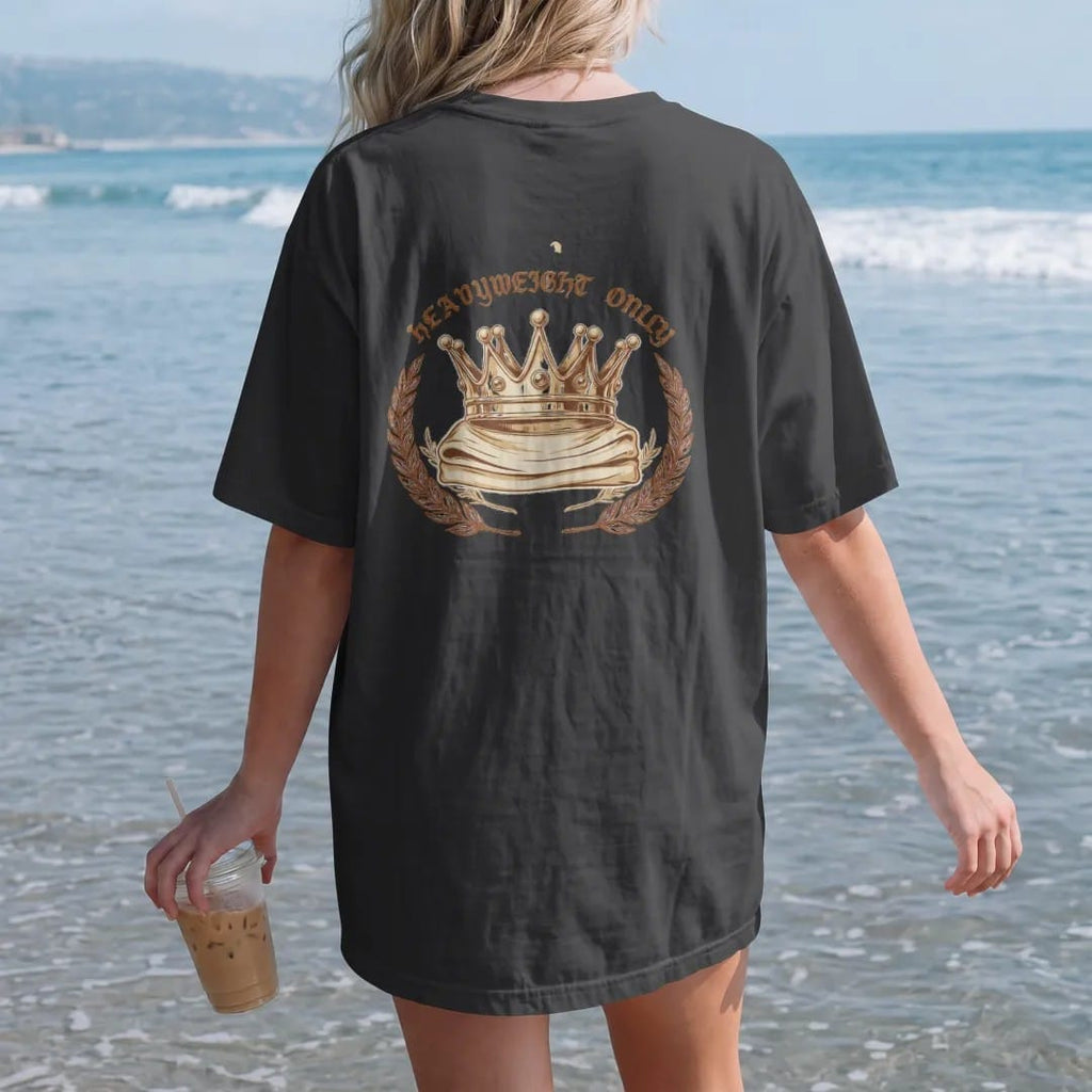Woman on the beach in a Black tee, back view: a jeweled gold crown resting on a folded heavy tee between laurel branches, Heavyweight Only in blackletter above.