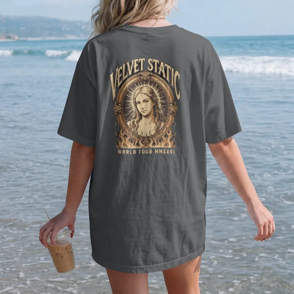 Woman on the beach in a Graphite tee, back view: a static-haloed angel in a cracked oval over flames, under Velvet Static in distressed blackletter and over World Tour MMXXVI in condensed capitals, in faded cream, silver and gold.