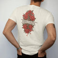 Man wearing a Ivory tee, seen from the back: roses and peonies blooming through a strand of barbed wire in engraved linework, with Printemps in fashion serif across the center, in espresso, oxblood and bronze.