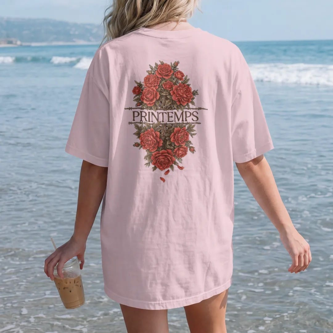 Woman on the beach in a Blossom tee, back view: roses and peonies blooming through a strand of barbed wire in engraved linework, with Printemps in fashion serif across the center, in espresso, oxblood and bronze.