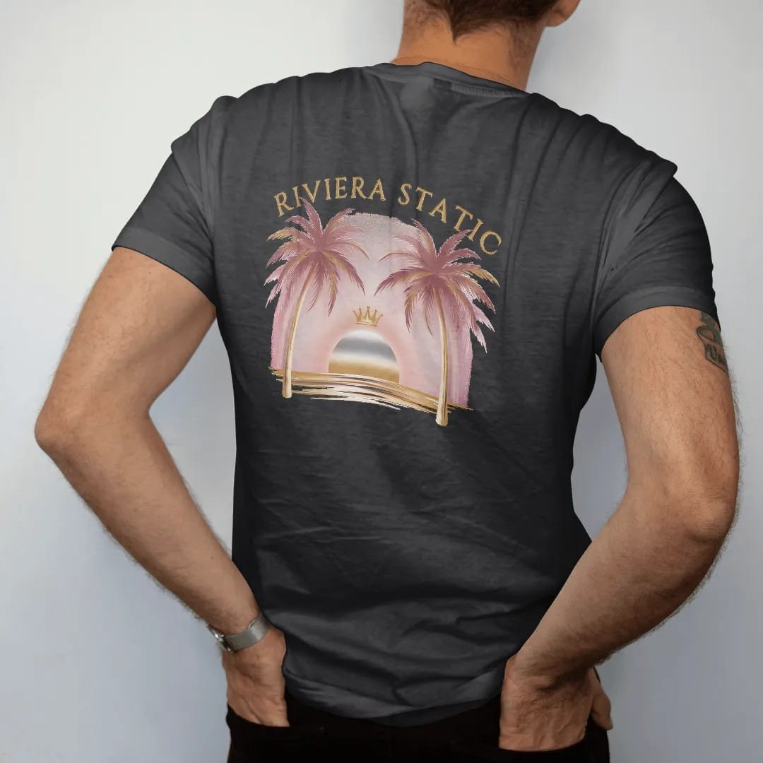 Man wearing a Black tee, seen from the back: an airbrush-style sunset over the sea between two palm trees with a small crown above the sun, under Riviera Static in arched fashion serif, in dusty rose, antique gold, bone cream and chrome silver.