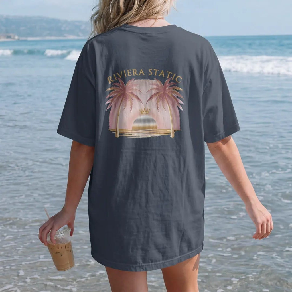 Woman on the beach in a Midnight tee, back view: an airbrush-style sunset over the sea between two palm trees with a small crown above the sun, under Riviera Static in arched fashion serif, in dusty rose, antique gold, bone cream and chrome silver.