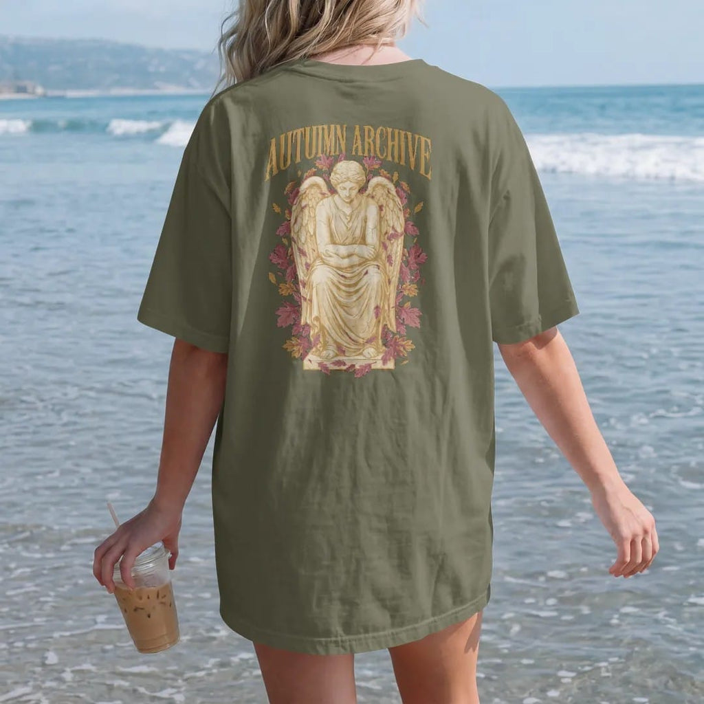 Woman on the beach in a Hemp tee, back view: a weathered marble angel seated with bowed head among falling oak and maple leaves, under Autumn Archive in gothic blackletter, in antique gold, dusty rose and bone cream.
