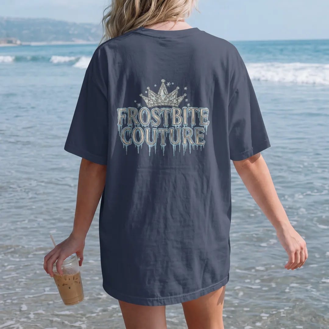 Woman on the beach in a Navy tee, back view: Frostbite Couture in tall frosted-chrome blackletter dripping icicles under a crown of ice crystals with rhinestone snow sparkles, in chrome silver, bone cream and ice blue.