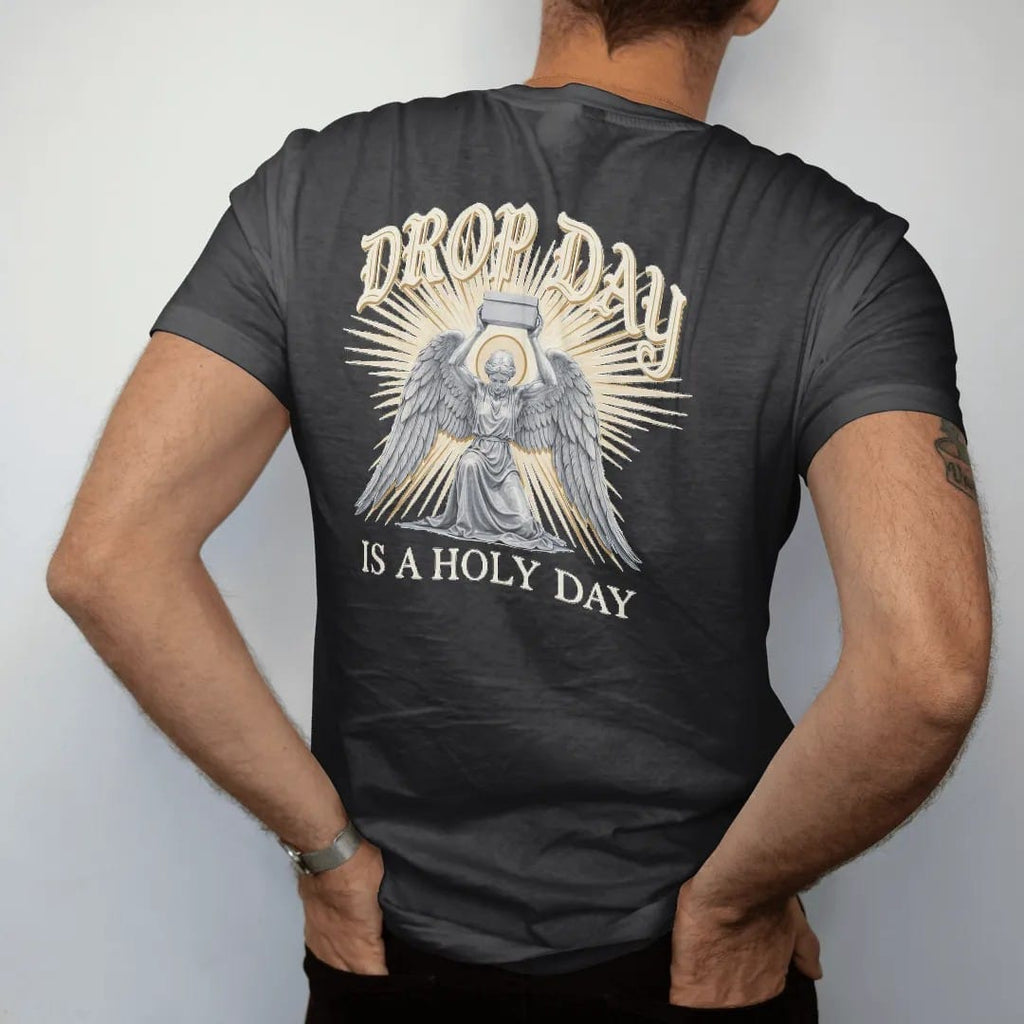 Man wearing a Black tee, seen from the back: a kneeling winged angel raising a plain sneaker box in beams of light, under Drop Day in blackletter and over Is A Holy Day in serif capitals, in bone cream, chrome silver and antique gold.