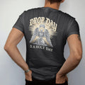 Man wearing a Black tee, seen from the back: a kneeling winged angel raising a plain sneaker box in beams of light, under Drop Day in blackletter and over Is A Holy Day in serif capitals, in bone cream, chrome silver and antique gold.