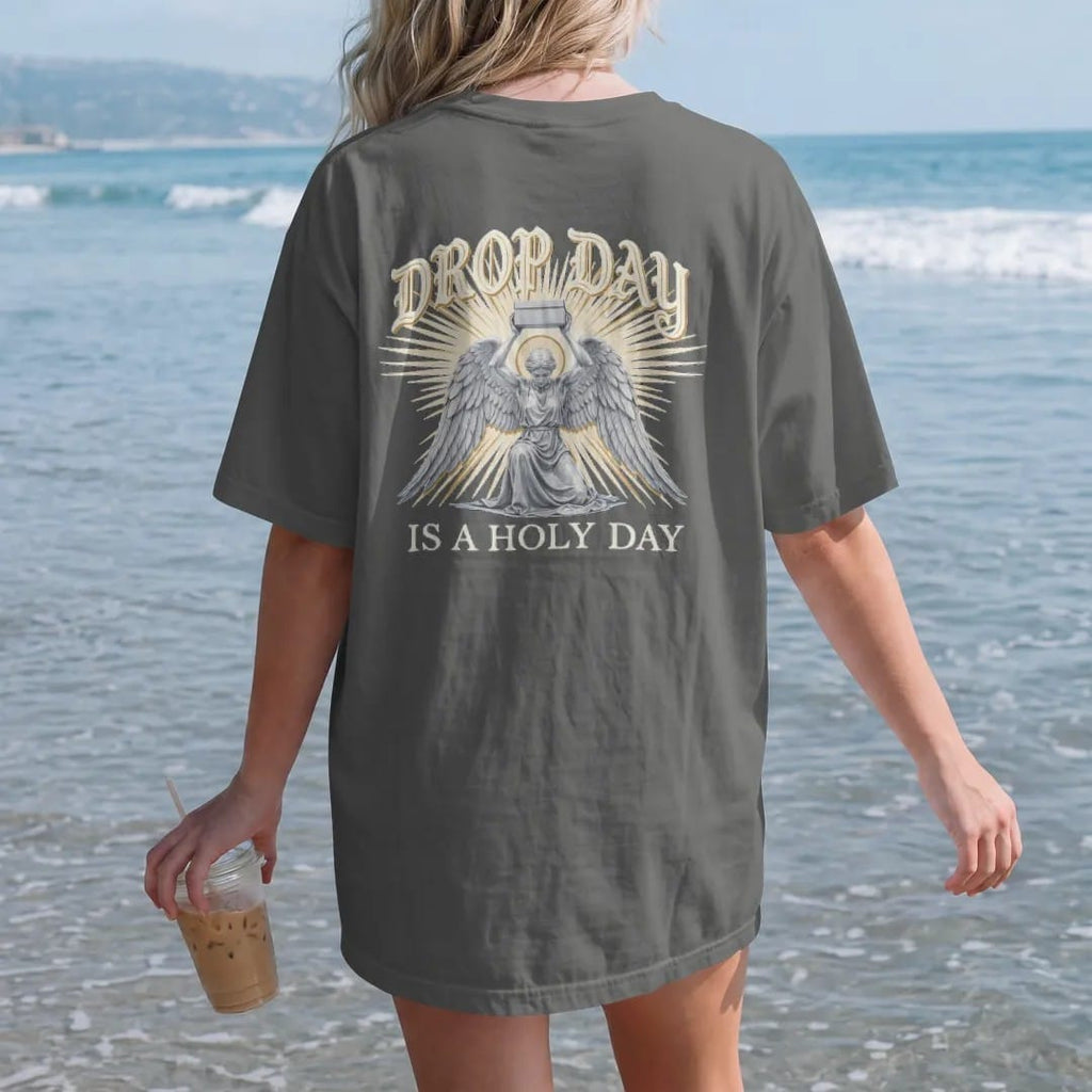 Woman on the beach in a Pepper tee, back view: a kneeling winged angel raising a plain sneaker box in beams of light, under Drop Day in blackletter and over Is A Holy Day in serif capitals, in bone cream, chrome silver and antique gold.
