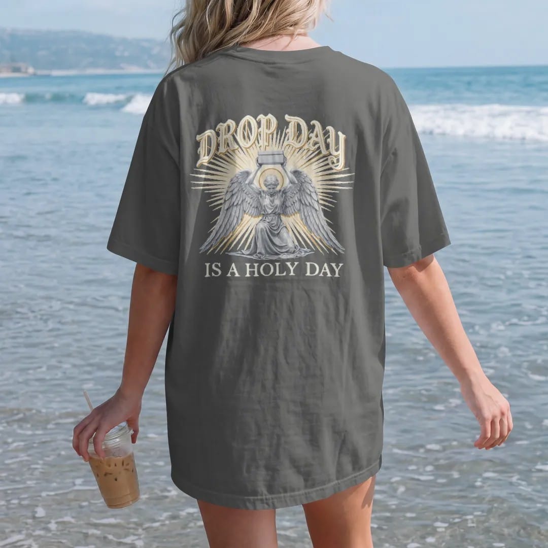 Woman on the beach in a Pepper tee, back view: a kneeling winged angel raising a plain sneaker box in beams of light, under Drop Day in blackletter and over Is A Holy Day in serif capitals, in bone cream, chrome silver and antique gold.