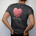 Man wearing a Black tee, seen from the back: a glossy airbrushed heart wrapped in barbed wire dissolving into static and glitch lines, above Love Is Static in gothic blackletter, in dusty rose, chrome silver and bone cream.