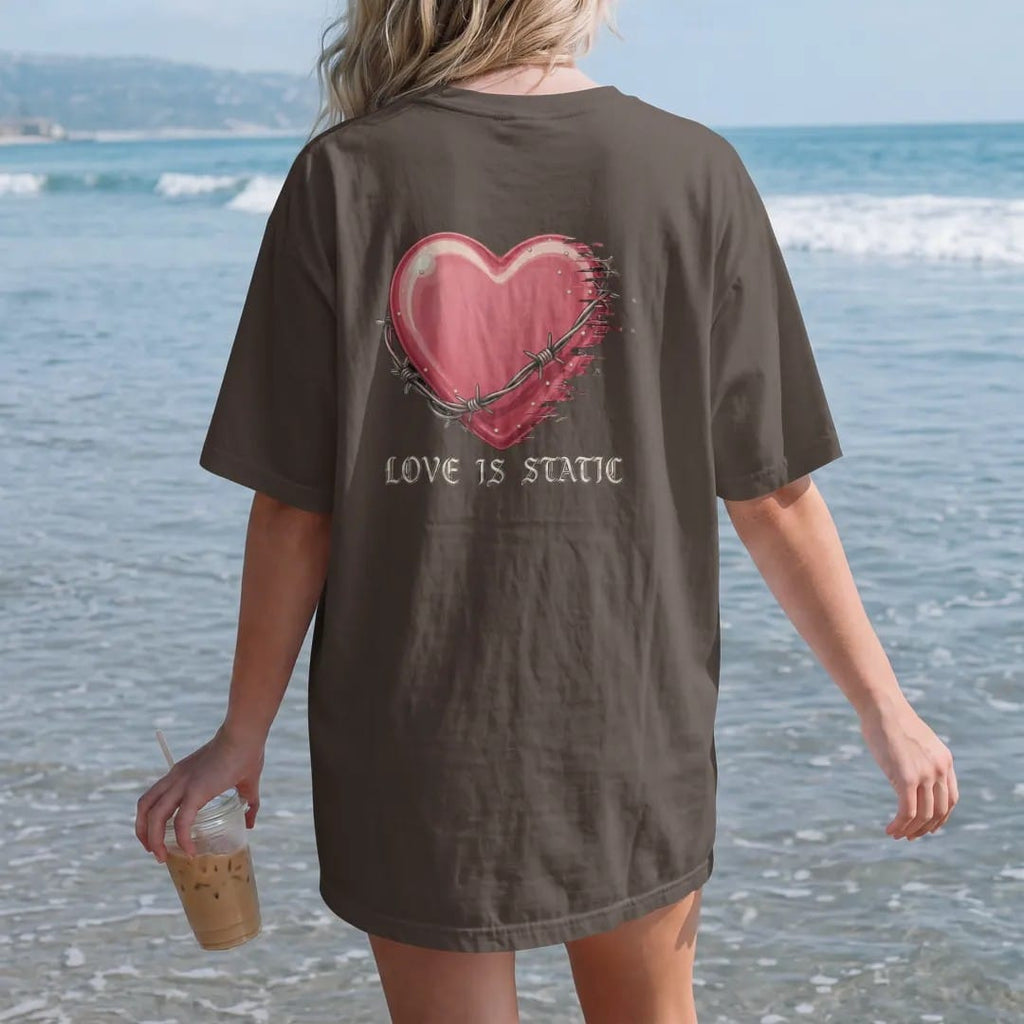 Woman on the beach in a Espresso tee, back view: a glossy airbrushed heart wrapped in barbed wire dissolving into static and glitch lines, above Love Is Static in gothic blackletter, in dusty rose, chrome silver and bone cream.