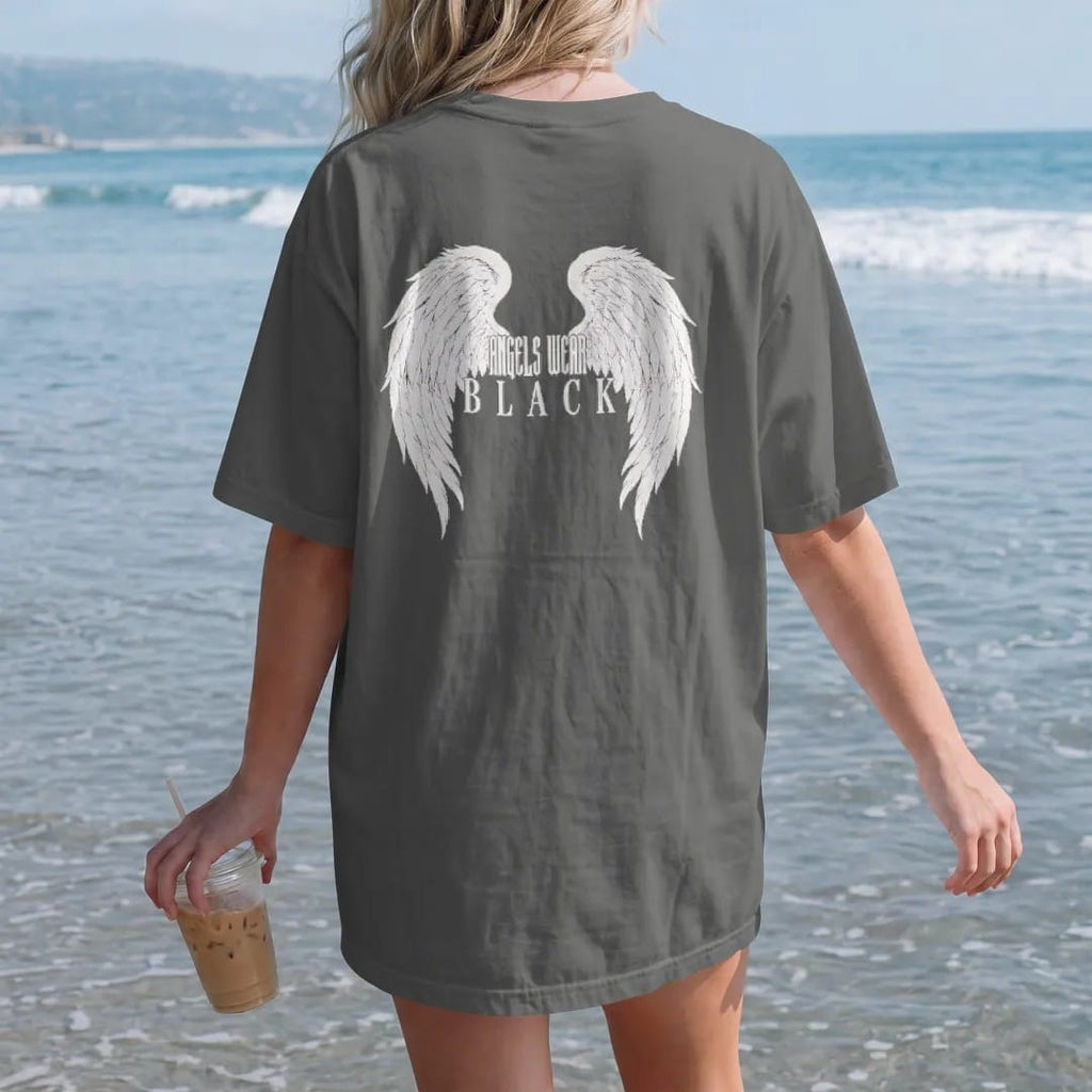 Woman on the beach in a Pepper tee, back view: a huge pair of spread engraved angel wings with Angels Wear in gothic blackletter between them and Black in spaced serif capitals beneath, in single-ink bone cream.
