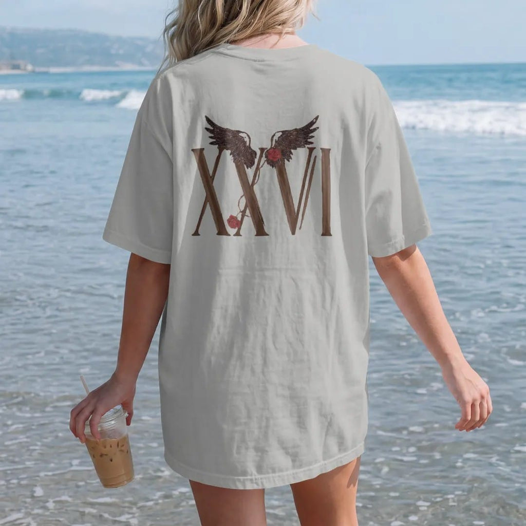 Woman on the beach in a Granite tee, back view: Tall Roman serif numerals XXVI with a pair of feathered wings above and a thorny rose vine, in espresso and oxblood.