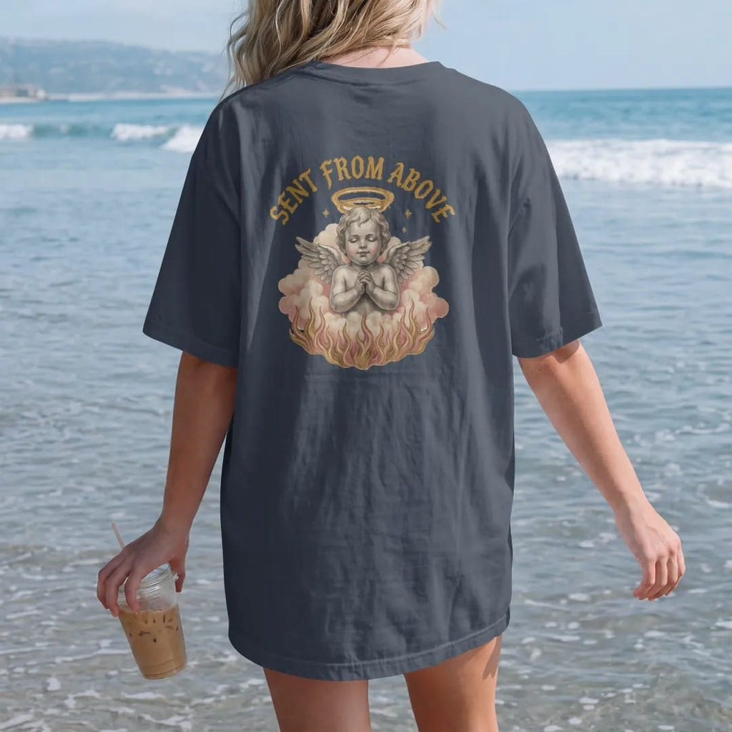 Woman on the beach in a Midnight tee, back view: a 90s airbrush-style cherub on clouds with a static halo and flames below, under Sent From Above in arched blackletter, in dusty rose, antique gold, chrome silver and bone cream.
