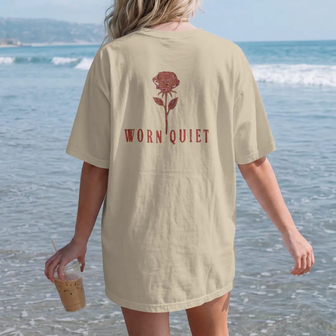 Woman on the beach in a Sandstone tee, back view: a single fine-line long-stemmed rose above Worn Quiet in large widely spaced didone serif capitals, in espresso and oxblood.