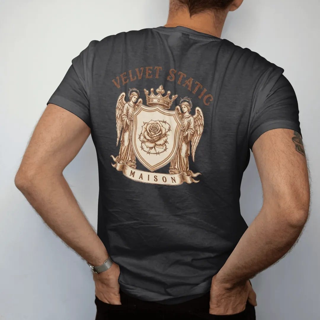 Man wearing a Black tee, seen from the back: a heraldic shield with a barbed-wire rose guarded by two static-haloed angels under a crown, with Velvet Static in arched blackletter and Maison on the ribbon, in bone cream, chrome silver and antique…