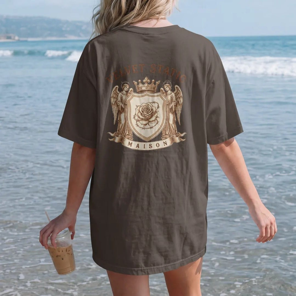 Woman on the beach in a Espresso tee, back view: a heraldic shield with a barbed-wire rose guarded by two static-haloed angels under a crown, with Velvet Static in arched blackletter and Maison on the ribbon, in bone cream, chrome silver and…