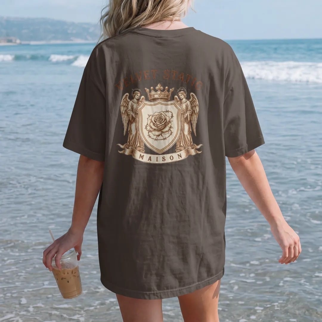 Woman on the beach in a Espresso tee, back view: a heraldic shield with a barbed-wire rose guarded by two static-haloed angels under a crown, with Velvet Static in arched blackletter and Maison on the ribbon, in bone cream, chrome silver and…