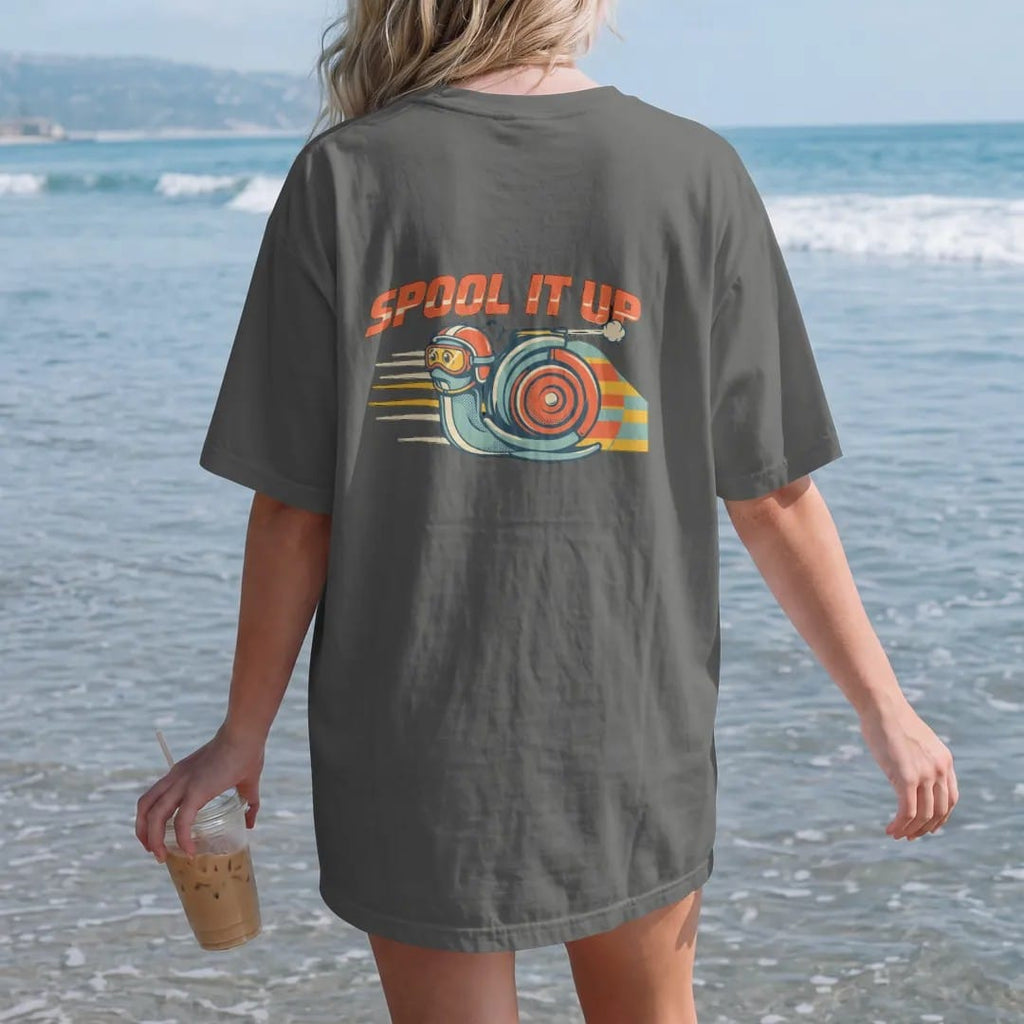 Woman on the beach in a Pepper tee, back view: a helmeted turbo snail mascot speeding over rally stripes under arched Spool It Up lettering, in cream, rally orange, signal yellow and teal.