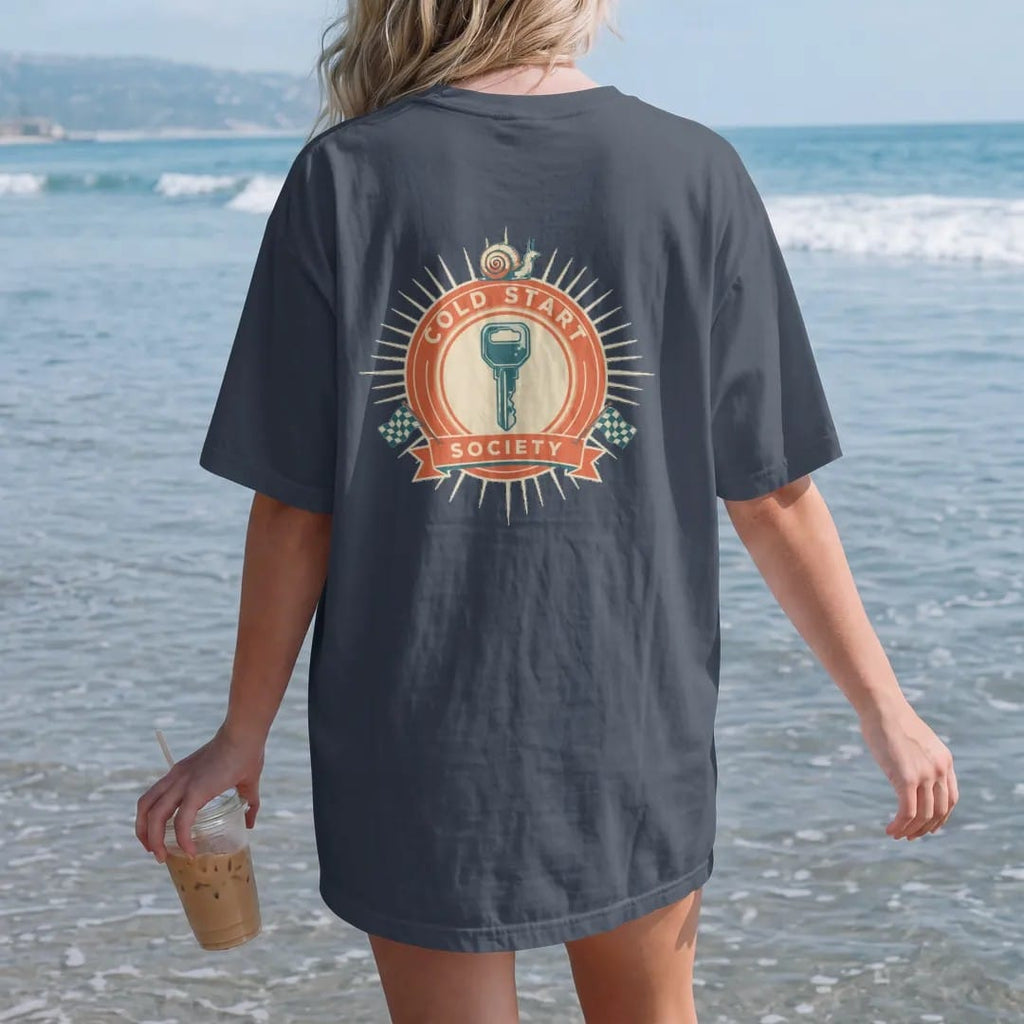 Woman on the beach in a Midnight tee, back view: a racing roundel with a frosted ignition key and a small turbo snail, Cold Start arched on top and Society on a bottom banner, in cream, orange, yellow and teal.