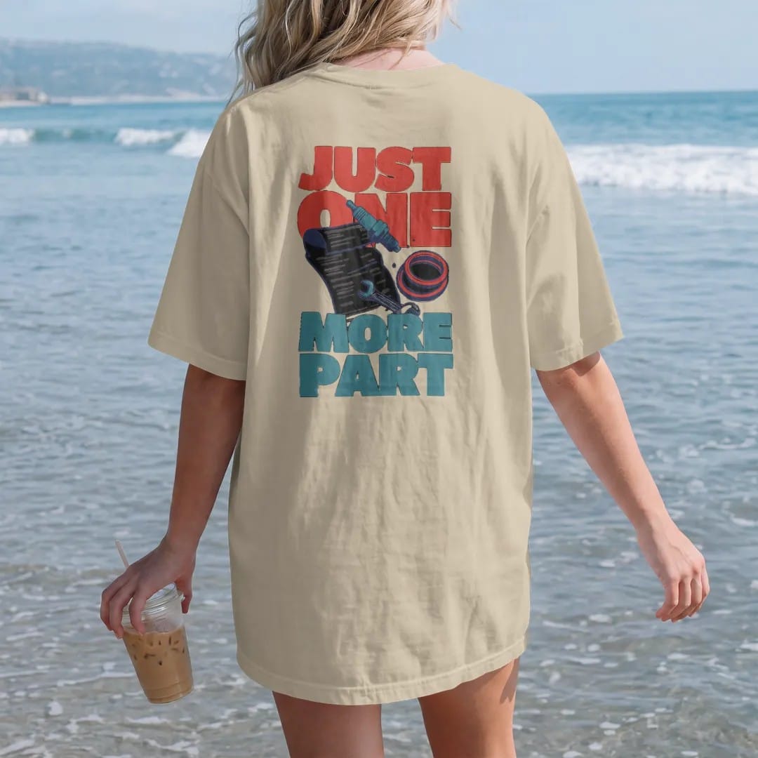 Woman on the beach in a Sandstone tee, back view: a long curling parts-store receipt spilling a spark plug, gasket and wrench, with Just One More Part in stacked letters, in charcoal, rally red, teal and navy.