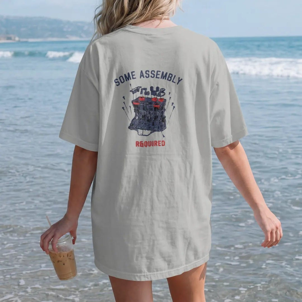 Woman on the beach in a Granite tee, back view: an exploded blueprint diagram of a generic inline engine with floating pistons, valves and bolts under Some Assembly Required, in navy, rally red and charcoal.