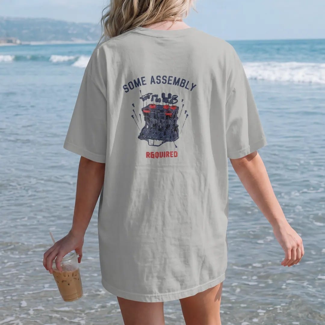 Woman on the beach in a Granite tee, back view: an exploded blueprint diagram of a generic inline engine with floating pistons, valves and bolts under Some Assembly Required, in navy, rally red and charcoal.