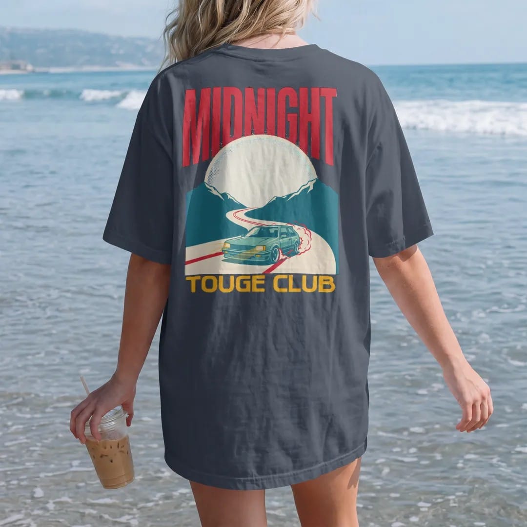 Woman on the beach in a Midnight tee, back view: a generic 90s hatchback drifting a moonlit mountain hairpin in a Japanese poster layout with Midnight Touge Club lettering, in cream, red, yellow and teal.