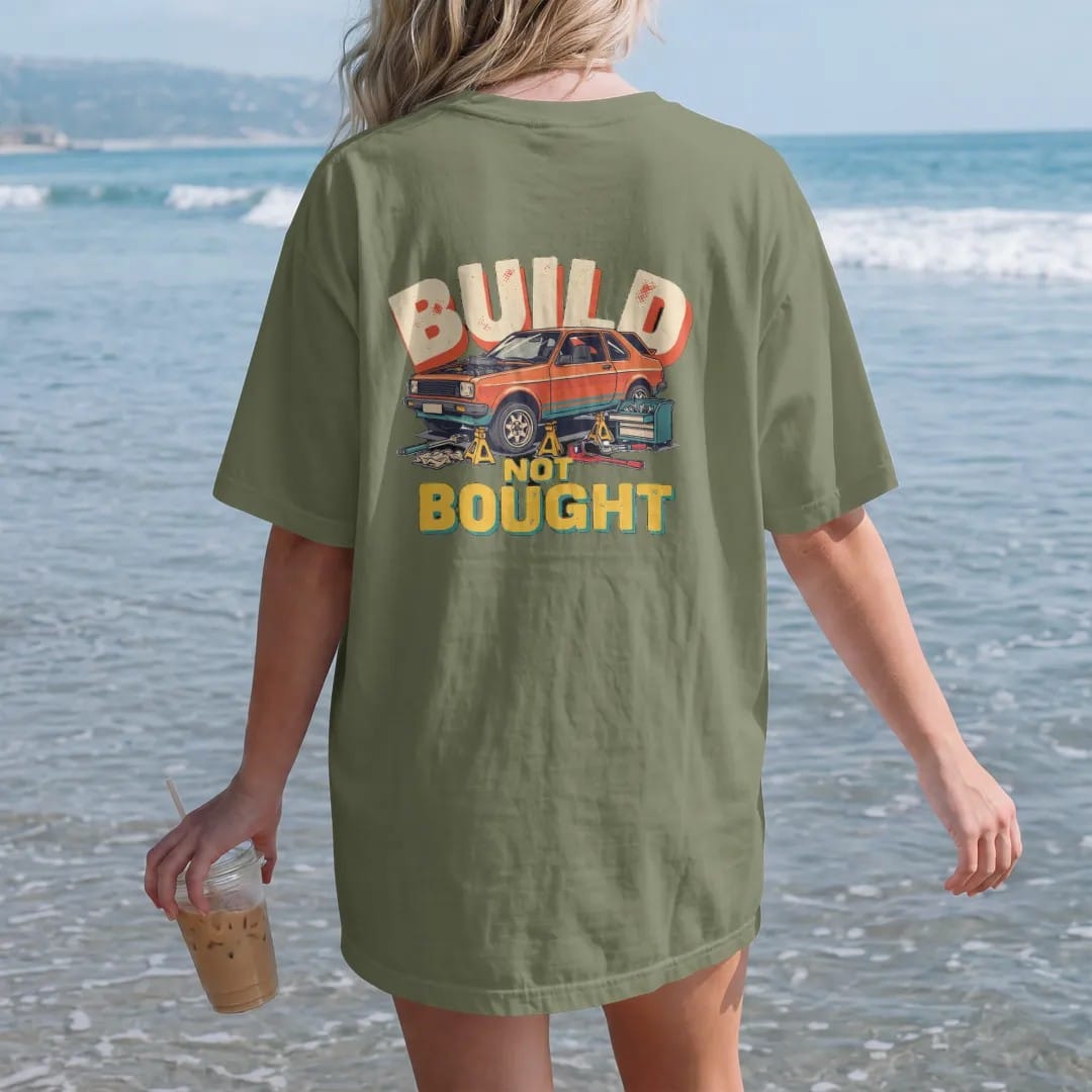 Woman on the beach in a Moss tee, back view: a generic project car up on jack stands with no wheels and the hood off, beside a creeper and toolbox, under Build Not Bought, in cream, orange, teal and yellow.