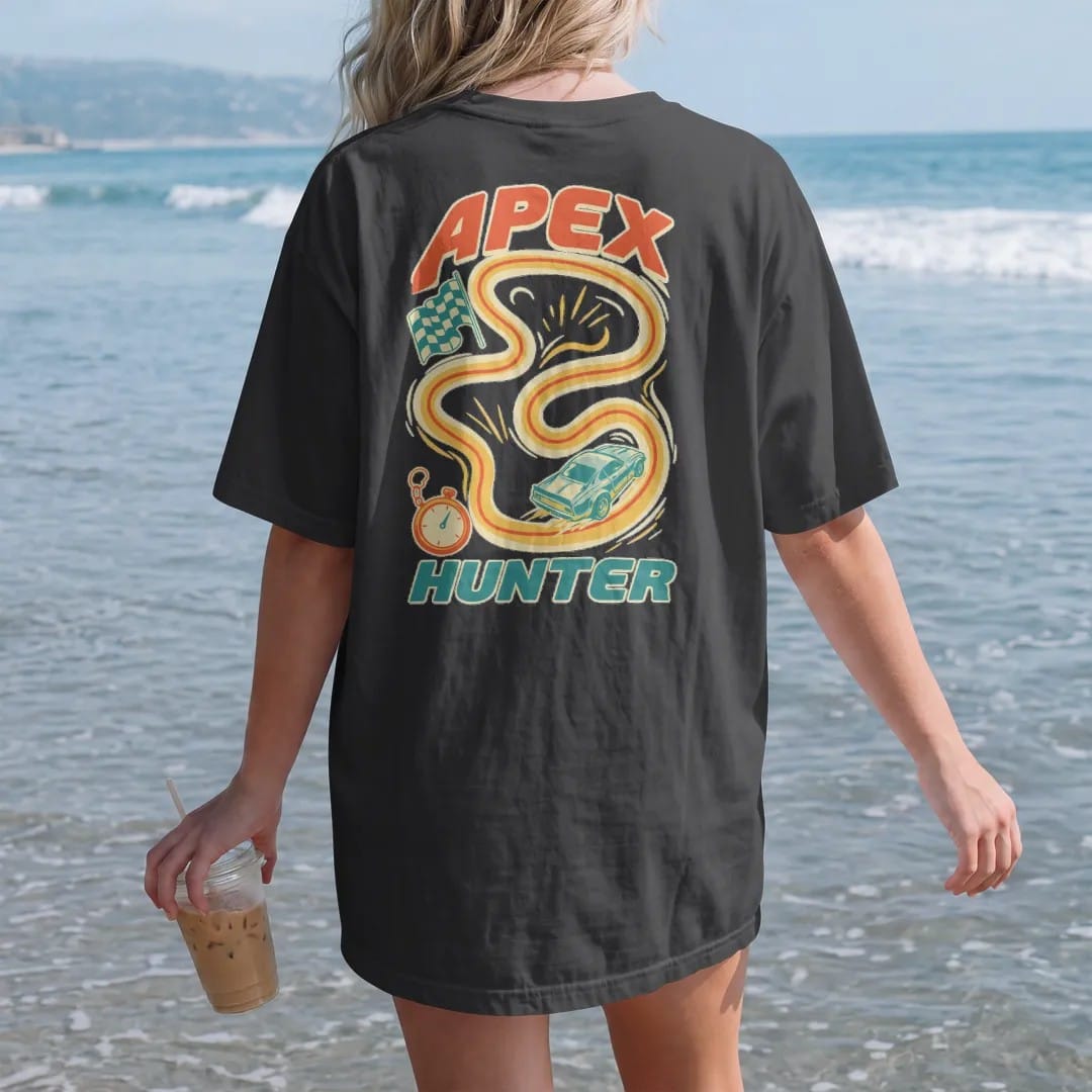 Woman on the beach in a Black tee, back view: a winding race-track map with a glowing racing line, checkered flag and a generic race car diving into a turn, with Apex Hunter lettering, in cream, orange, teal and yellow.