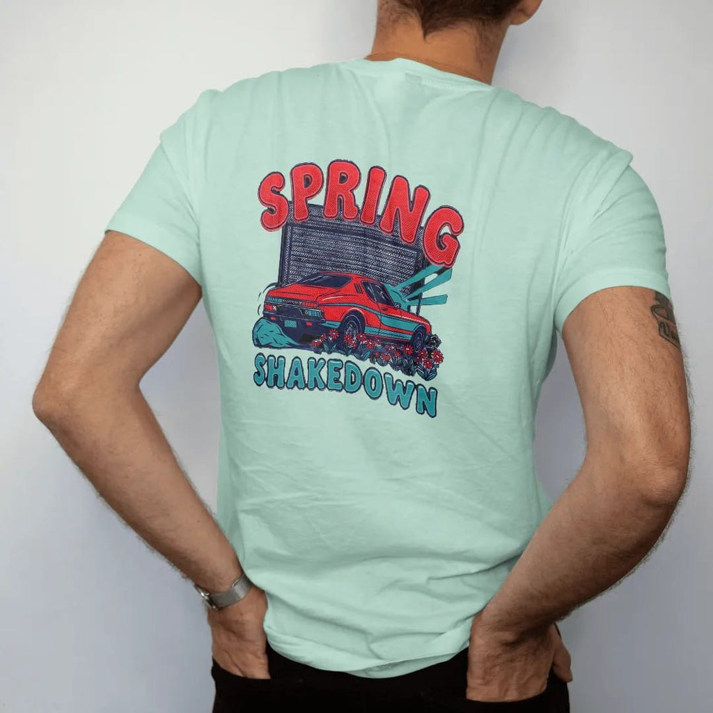 Man wearing a Chalky Mint tee, seen from the back: a generic classic coupe rolling out of an open garage into spring sun with daisies by the tires, Spring Shakedown lettering, in charcoal, rally red, teal and navy.