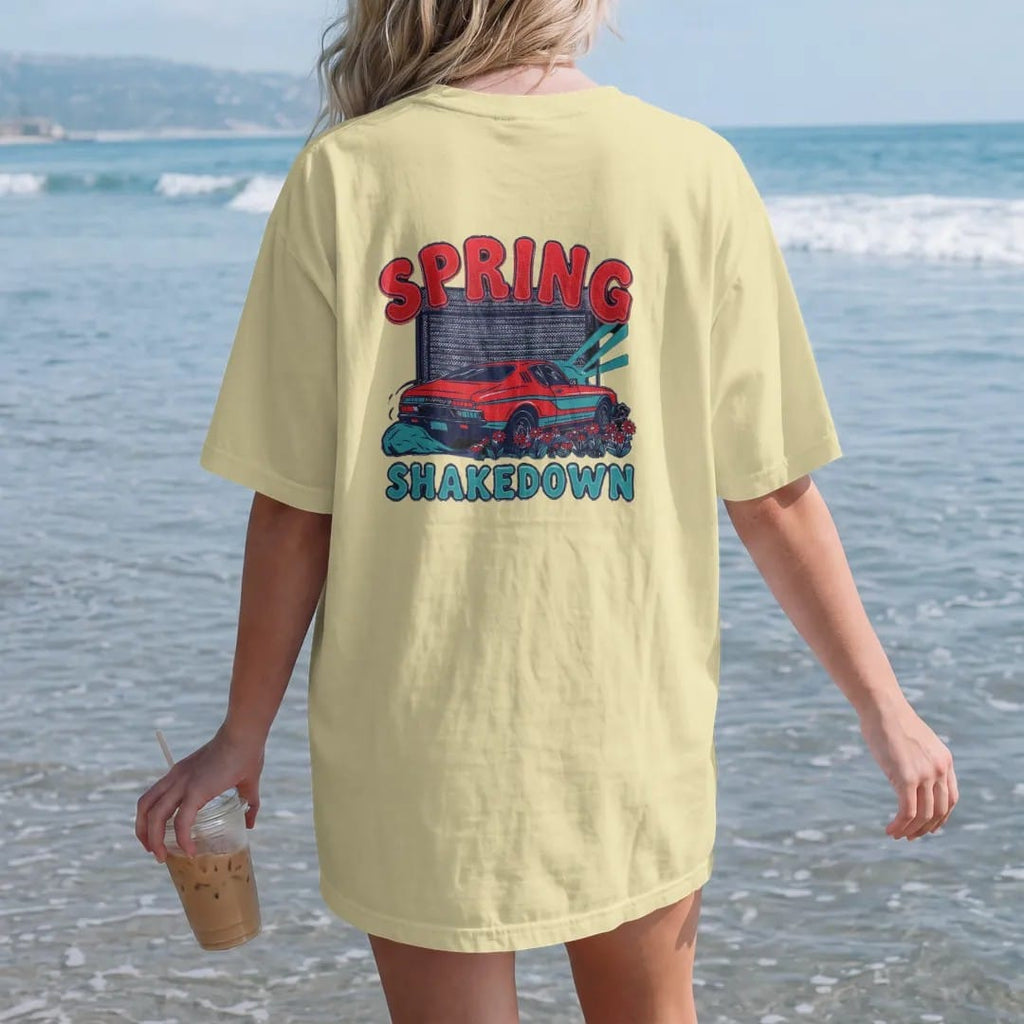 Woman on the beach in a Butter tee, back view: a generic classic coupe rolling out of an open garage into spring sun with daisies by the tires, Spring Shakedown lettering, in charcoal, rally red, teal and navy.