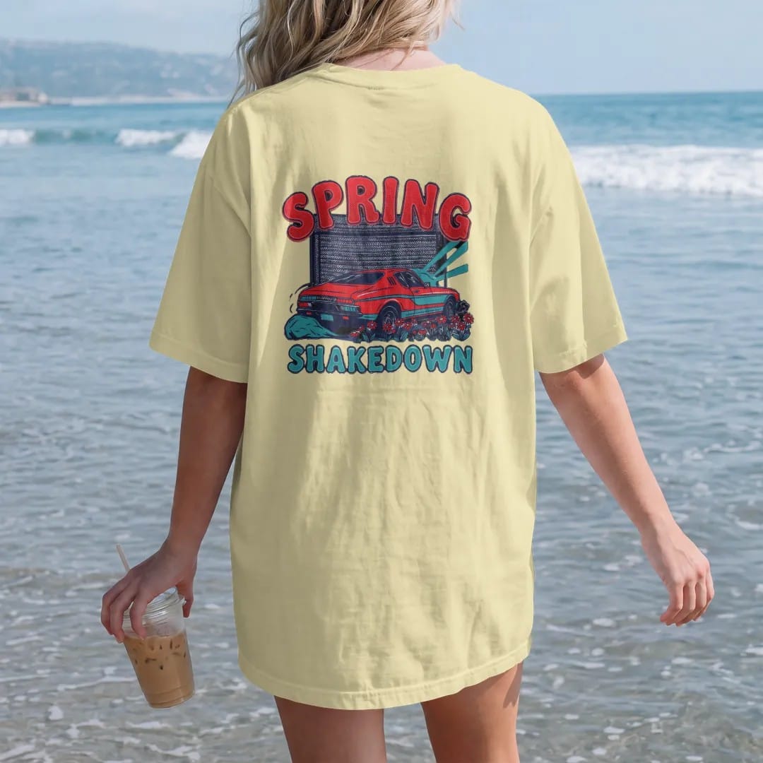 Woman on the beach in a Butter tee, back view: a generic classic coupe rolling out of an open garage into spring sun with daisies by the tires, Spring Shakedown lettering, in charcoal, rally red, teal and navy.