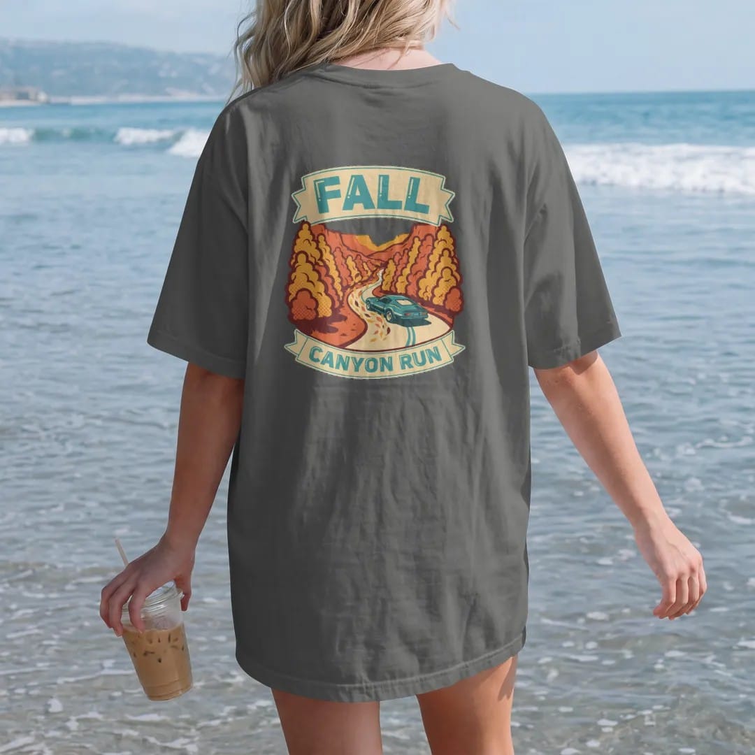 Woman on the beach in a Pepper tee, back view: a generic sports coupe carving a canyon road through orange and gold trees with swirling leaves, Fall Canyon Run lettering, in cream, burnt orange, yellow and teal.