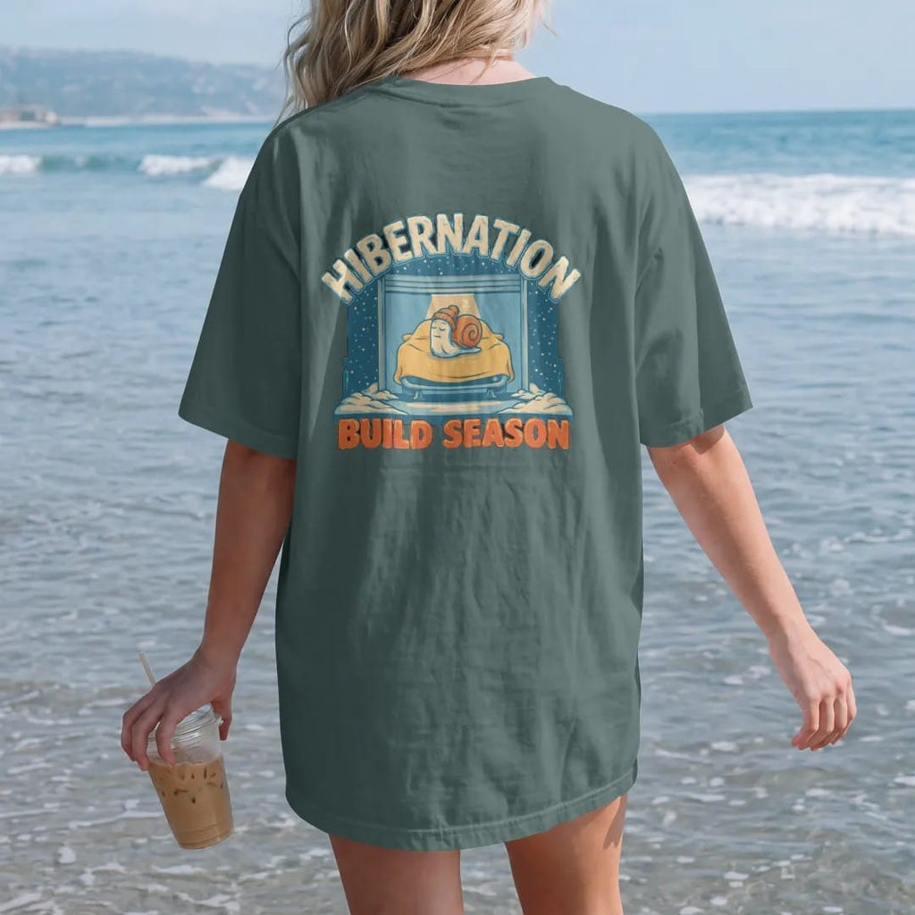 Woman on the beach in a Blue Spruce tee, back view: a snowy garage with a glowing window, a covered generic car on jack stands and the turbo snail asleep on the hood, Hibernation Build Season lettering, in cream, icy teal, yellow and orange.